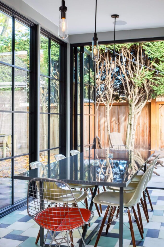 A modern dining area with a glass table, white chairs, and one wire chair with an orange cushion. Large windows offer a view of a fenced backyard with trees and patio chairs. Geometric tile flooring is visible.
