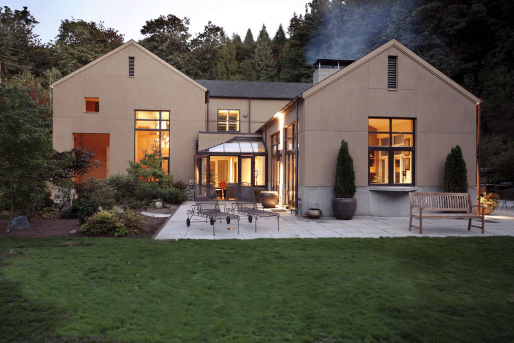 Modern house with large windows and warm interior lighting, featuring two main sections connected by a glass entryway. The backyard has a patio with metal furniture, potted plants, a wooden bench, and is surrounded by trees and grass.
