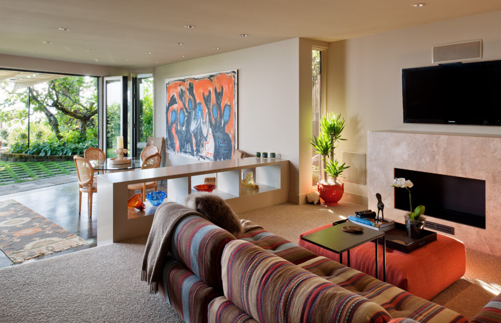 A modern living room with a striped sofa, a wall-mounted TV above a fireplace, abstract artwork on the wall, and large windows opening to a patio with greenery outside.