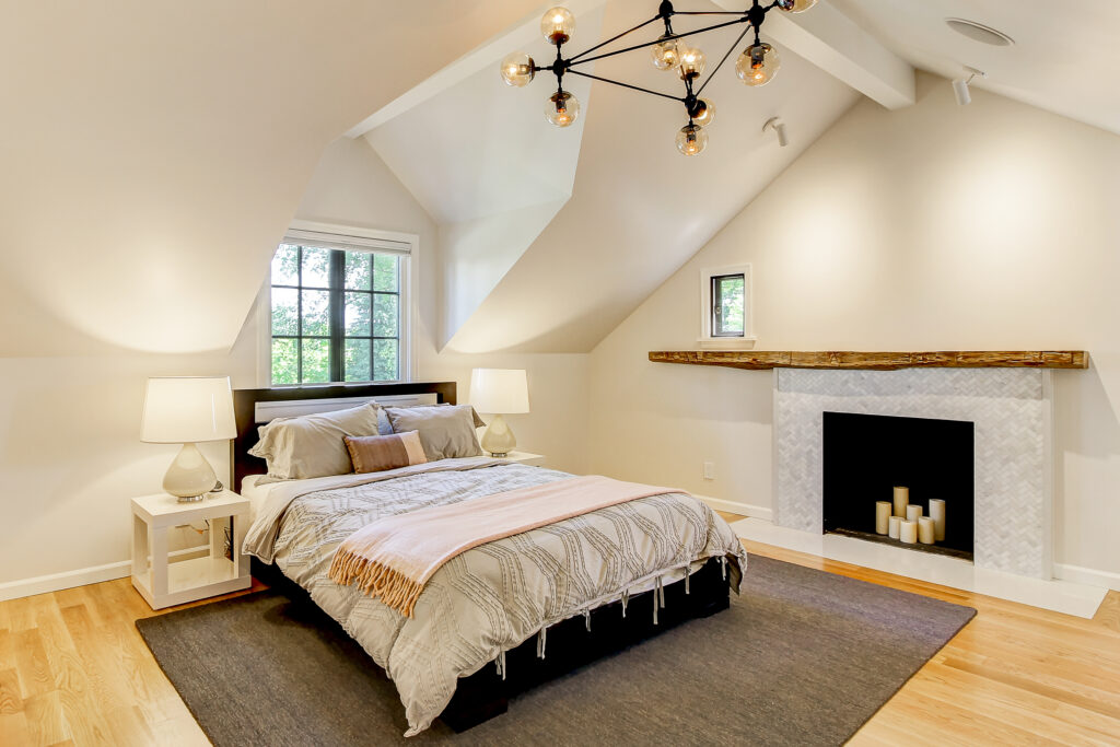 A cozy bedroom with sloped ceilings, a bed with gray and beige bedding, two bedside tables with lamps, a modern chandelier, a fireplace with candles, hardwood floors, and a window letting in natural light.