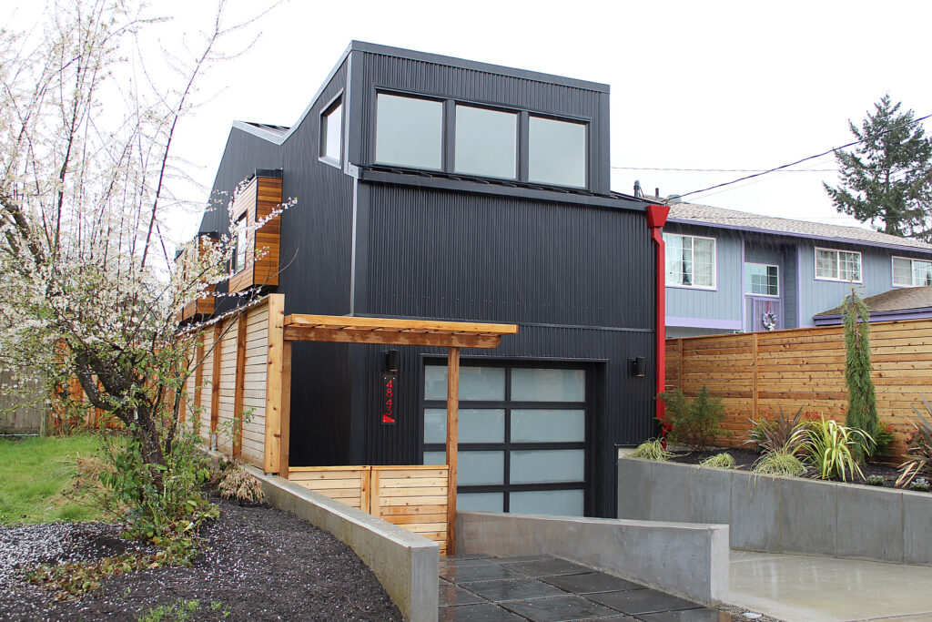 A modern black house with large windows and a wood accent, featuring a glass garage door and a small wooden pergola at the entrance. The yard has new landscaping and a concrete driveway.