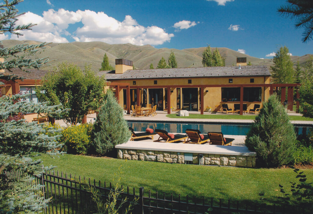 A modern house with a pool and lounge chairs sits in front of rolling hills under a blue sky with white clouds.