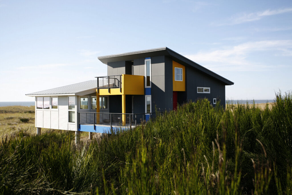 A modern two-story house with gray, white, yellow, and orange accents stands in a grassy, coastal landscape under a blue sky. The house has large windows, a balcony, and angular rooflines.