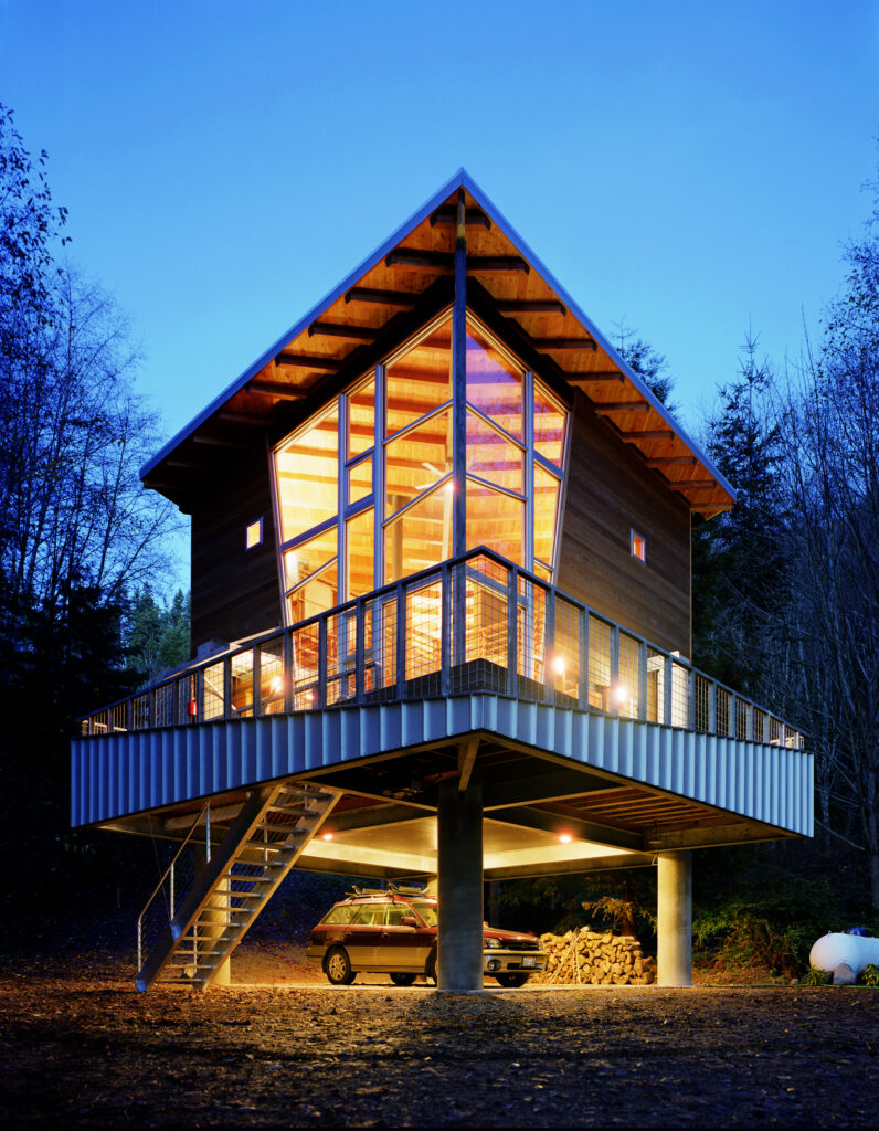 A modern house with large glass windows and wooden panels stands elevated on stilts above the ground. An outdoor staircase leads up to the entrance, and a car is parked underneath the house among trees at dusk.