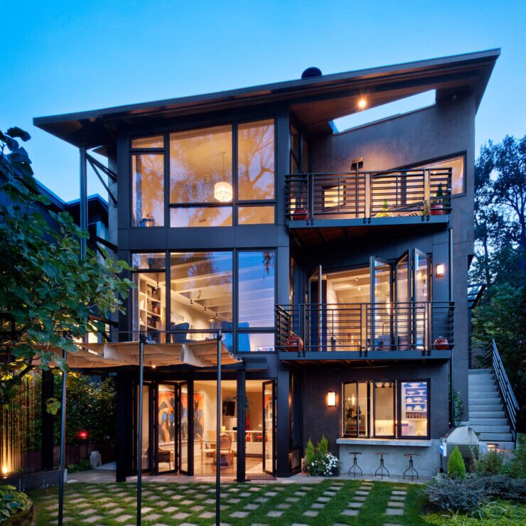 A modern three-story house with large glass windows, multiple balconies, outdoor seating, and warm interior lighting, surrounded by greenery at dusk.