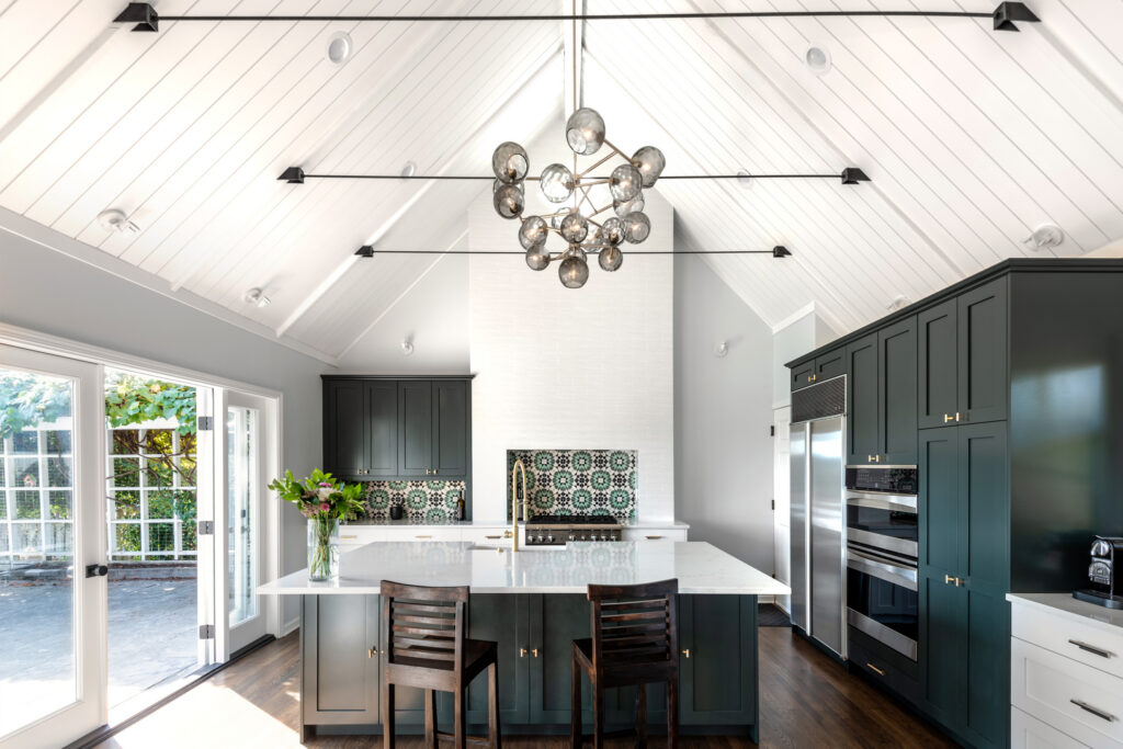 Modern kitchen with vaulted white ceiling, dark green cabinets, a large island with two chairs, floral tile backsplash, and a round glass chandelier. Sunlight streams in through large glass doors on the left.
